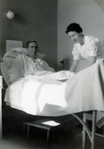 Ett kvinnligt sjukvårdsbiträde står bredvid en manlig patient som ligger i en sjuksäng på den manliga kirurgavdelningen, Mölndals sjukhus 1939 - 1941. Baktill på sänggaveln sitter en temperaturkurva. Bilder från fotoalbum, tagna från södra sidan av Länsmansgatan vid Mölndals lasarett/sjukhus 1939 - 1941 på bland annat givarens mor Ester (född Johansson) tillsammans med kollegan/väninnan Aina Englund. De arbetade på den manliga kirurgavdelningen som vårdbiträden/undersköterskor.
Relaterade motiv: 2024_1542 - 1617.