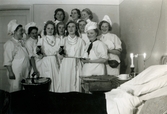 Elva lusseklädda kvinnor, sju sjukvårdsbiträden och fyra kökspersonal med vita kockmössor, står samlade bredvid en tom sjuksäng på den manliga kirurgavdelningen på Mölndals sjukhus 1939 - 1941. På sängbordet finns två tända ljus och ett par kaffekoppar. Baktill på sänggaveln sitter en temperaturkurva. Bilder från fotoalbum, tagna från södra sidan av Länsmansgatan vid Mölndals lasarett/sjukhus 1939 - 1941 på bland annat givarens mor Ester (född Johansson) tillsammans med kollegan/väninnan Aina Englund. De arbetade på den manliga kirurgavdelningen som vårdbiträden/undersköterskor.
Relaterade motiv: 2024_1542 - 1617.