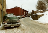 Parkerade bilar på vänster sida av Royens gata intill industribyggnader. Kvarnbyn i Mölndal, januari 1984.