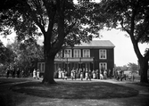 Okänd större mangårdsbyggnad med balkong, ca 1912. En stor samling människor står framför huset.