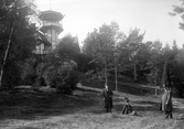 Två män och en kvinna i en park vid en högt belägen villa med torn. Okänd plats, ca. 1910-tal.