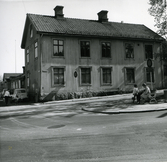 Skräddarmästare John Edqvists gård i hörnet av Stora Kyrkogatan och Repslagargatan, kvarteret Kyrkbacken 2, Hudiksvall. På bilden syns en tvåvåningsbyggnad med tegeltak och två skorstenar. Till höger syns en parkbänk med ett par människor. Till vänster gående människor samt parkerad bil utmed husväggen.