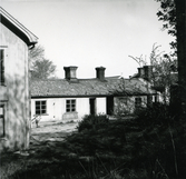 Envåningslängan mot Repslagargatan, gårdssidan, i kvarteret Kyrkbacken 2, Hudiksvall. Till vänster syns gaveln på en byggnad i två våningar. I förgrunden anas träd, buskar och gräs.