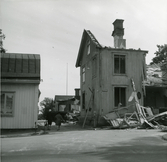 Rivningshus i kvarteret Kyrkbacken 2, Hudiksvall. En gavel och skorstenen står fortfarande kvar. Till vänster syns prästgården. Två personer går mellan byggnaderna.