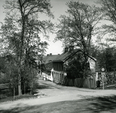 Till höger syns en trafikskylt och ett staket, därefter skräddare Edqvists gård vid Stora Kyrkogatan, kvarteret Kyrkbacken, Hudiksvall. Längre ner i backen Prästgården.