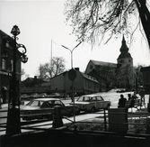 Vy över kvarteret Prästgården och bortanför det kvarteret Kyrkbacken 2, sett från kanalen. Närmast står två bilar parkerade och några människor sitter på en parkbänk till höger. Till vänster en gatubelysning med tre lyktor. I bakgrunden syns kyrkan.