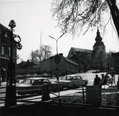 Vy över kvarteret Prästgården och bortanför det kvarteret Kyrkbacken 2, sett från kanalen. Närmast står två bilar parkerade och några människor sitter på en parkbänk till höger. Till vänster en gatubelysning med tre lyktor. I bakgrunden syns kyrkan.