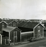 Två av sjöbodarna vid Magasinsgatan, kvarteret Köpmannen, Hudiksvall. Vid en av dörrarna står ett antal lådor/backar och en bil står parkerad utmed väggen. I bakgrunden syns sjöbodarna på andra sidan Sundskanalen. Till höger anas Östra Skolan.