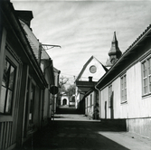 Lilla Kyrkogatan, Hudiksvall, före rivning av kvarteret Kyrkbacken. Till höger församlingssalen i kvarteret Prästgården. På vänster sida hotell Fridhem. I bakgrunden en av kyrkportalerna, kyrkans gavel samt kyrktornet.