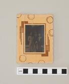 Fotografi inramat i ram av papp. Föreställande tre personer. En kvinna i ljus dräkt, halvt uppsatt hår och hållandes en tunn kappa eller möjligen sjal. Längst bak en glasögonprydd man med hatt, kostym och slips. I förgrunden en yngling iförd kostym, ljus skjorta som är uppknöppt i Hållandes jacka/kappa samt nåon typ av platt väska/portfölj i armvecket.