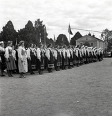 Jubileumsutställningen. Invigningen. 
En stor samling kvinnor i folkdräkt (Värendsdräkt m.m.) har
ställt upp sig på gårdsplanen framför Växjö Högre Allmänna Läroverk. 
I bakgrunden skymtar de gamla flyglarna till Växjö lasarett.