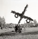 En midsommarstång håller på att resas på en äng. 
I bakgrunden skymtar delar av ett tivoli.