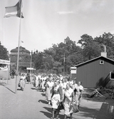 Ett antal kvinnor i husmorsdräkter är på väg genom utställningsområdet.
I bakgrunden ser man taket m.m. på dåv. Växjö Högre Allmänna Läroverk.