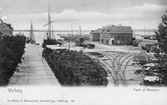 Två kort av samma fotografi med vy från Södra Hamnvägen över hamnområdet i Varberg med magasin och räls för godsvagnar. I bakgrunden ligger Getterön. Troligen från ca 1905-1910.  
Bild 1: Brevkort, 