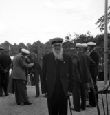 En äldre, skäggig man i studentmössa m.m., poserar för fotografen framför
Växjö Högre Allmänna Läroverk. I bakgrunden diskuterar några andra f.d. studenter något. Växjöstudenternas dag.