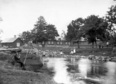 Gård vid en å. Vid åkanterna befinner sig ett antal människor. Okänd plats, tidigt 1900-tal.