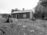 Kvinna på en sten framför en stuga. Okänd plats, tidigt 1900-tal.