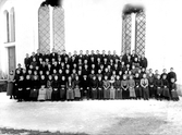 Konfirmander fotograferade utanför kyrka. I mitten sitter prästen. Tidigt 1900-tal.