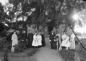 Bröllopsfölje uppställda framför hus, möjligen lanthandel eller bostadshus. Ca. 1910.