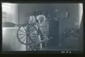 En äldre kvinna arbetar vid en spinnrock i ett rum.

