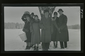 Grupporträtt av några människor fotograferade utomhus i snön. Ett par bär hinkar och en man ett ris som det sitter en liten docka i.
