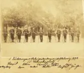 En grupp militärer med cyklar står uppställda på en grusplan.
I bakgrunden skymtar några hyddor.
Text under bilden: 