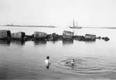 Två badande kvinnor vid Barnens badstrand. I bakgrunden Skrivarklippan med fyren, ett ång- och ett segelfartyg.