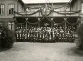 Gruppfoto av en stor samling officerare och civila herrar i frack och andra ämbetsuniformer, avfotograferade framför den festsmyckade officersmässen på Kronobergs hed. 
Källa: Svensk Rikskalender 1908,s. 173.