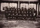 Gruppfoto av ett antal meniga/rekryter, uppställda utomhus. 
I mitten sitter några officerare. Till höger syns en regementsmusiker med diverse olika instrument.