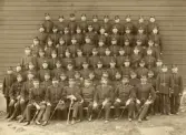 Gruppfoto av ett antal meniga/rekryter, uppställda utomhus, framför en rödmålad byggnad.
I mitten sitter några officerare.