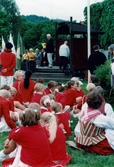 Nationaldagsfirande i Stadshusparken i Mölndal den 6 juni 1999. I bakgrunden ses en man hålla ett tal på scenen. I förgrunden sitter medlemmar ur Mölndals Paradorkester i gräset.