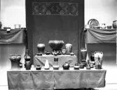 Utställning av keramikföremål, vaser, skålar, krukor och fat, i bakgrunden vävnad med bård i rölakan.