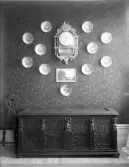 Interiör, snidad kista och stol samt väggur, tavla och tallrikar.