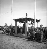 Kamratmötet, I 11. Samling på övningsfältet. En man i uniform (m/23), håller tal från den girlandprydda tribunen.