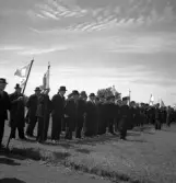 Kamratmötet, I 11. Samling på övningsfältet. Ett antal äldre herrar i kostym och hatt m.m., har ställt upp sig med kompanifanor.