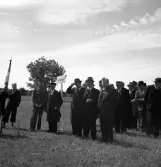 Kamratmötet, I 11. Samling på övningsfältet. Ett antal äldre herrar i kostym och hatt m.m., har ställt upp sig med kompanifanor m.m. En av herrarna pratar med en herre i uniform.