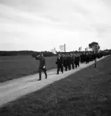 Kamratmötet, I 11. Ett antal äldre civilklädda herrar tågar fram på en liten grusväg med sina kompanifanor, med en herre i uniform (m/23) i täten.