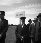 Kamratmötet, I 11. Några äldre herrar i kostym och hatt, med kompaniskylt, Livkompaniet. Till vänster syns en herre i uniform.