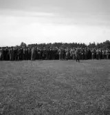 Kamratmötet, I 11. Äldre herrar i kostym och hatt, står uppställda på övningsfältet.