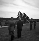 Kamratmötet, I 11. Övningsfältet. Förbidefilering av inryckt manskap i uniform. I förgrunden står några mannar med regementets fanor, jämte två andra herrar.