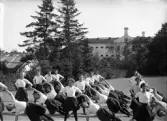 En trupp kvinnliga gymnaster övar i det fria utanför dåv. seminariet, dvs numera Ringsbergsskolan i Växjö.  I bakgrunden syns skolans växthus och dåv. cellfängelset.
