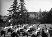 En trupp kvinnliga gymnaster övar i det fria utanför dåv. seminariet, dvs numera Ringsbergsskolan i Växjö.  I bakgrunden syns skolans växthus och  dåv. cellfängelset.