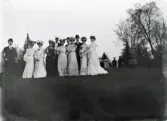 En samling damer i hatt m.m. tillsammans med två herrar, i någon slags parkanläggning.
Suddigt foto.