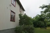 Bostadshusets baksida med dess vita träfasad och tre fönster med roströda karmar (se 2024_1535), Krokslättsgatan 18 i Mölndal 2023-06-21. Till höger ses växtlighet och trädgårdsmöbler i trädgården. Fotodokumentation av ett friliggande bostadshus/villa byggt 1932 med fyra (4) våningsplan. Byggnaden har tvättstuga i källaren samt torkvind på övervåningen. Boarea: 140 kvm, totalarea: 1004 kvm. Villan är ombyggd och fördelad på fyra (4) hyreslägenheter.
Relaterade motiv: 2024_1415 - 1541.
