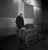 Avslutning vid Grimslövs folkhögskola.
En äldre man i glasögon och kostym, talar vid katedern/talarstolen i
gymnastiksalen.