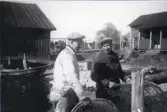Fiskarsönerna Arne Sjölin och Helge Söderhäll med en ryssja vid strandkant med sjöbod och stuga i bakgrunden, 1950-tal.