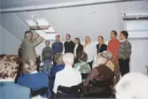 Körledaren Jan Ivar Blixt dirigerar en sångkör på åtta personer som sjunger framför en sittande publik (medlemmar) vid SVs årsmöte (Studieförbundet Vuxenskolan), ateljé Takåsen på Brogatan 16 i Mölndal cirka 2000. Körledaren har precis innan fått ett kulturpris.
Relaterade motiv: A4515 - A4518.
