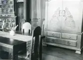 Rumsinteriör med bord och stolar, stort skåp och möjligen en kakelugn till vänster i bild, bordslöpare mm. troligen från Centralpalatset i Gävle stad.