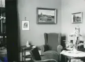 Rumsinteriör med fåtölj, golvlampa, tavlor, fotografi, sidobord, draperi mm. troligen från Centralpalatset i Gävle stad.