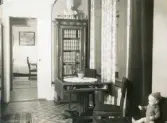 Rumsinteriör med rustikt bord och stolar, bokhylla med glasdörrar och stor byst högst upp, docka med kläder sitter till höger i bild, gardiner mm. troligen från Centralpalatset i Gävle stad.